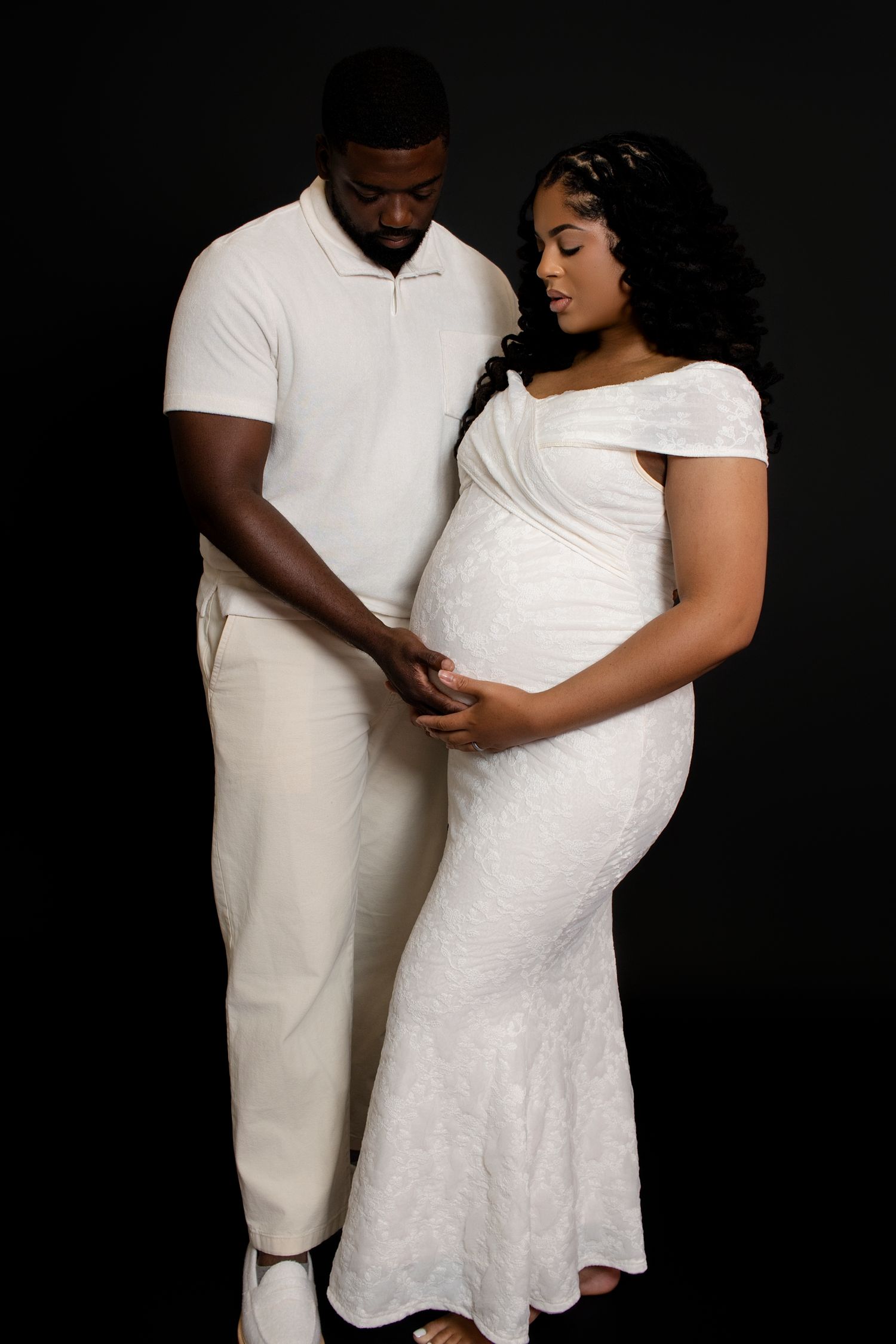 Intimate maternity portraits with couple wearing white against dark studio background.