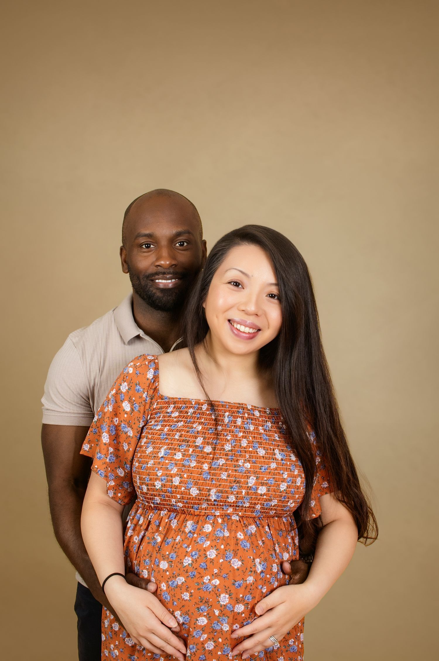 Maternity photo showing couple in orange floral dress and grey polo against beige background.