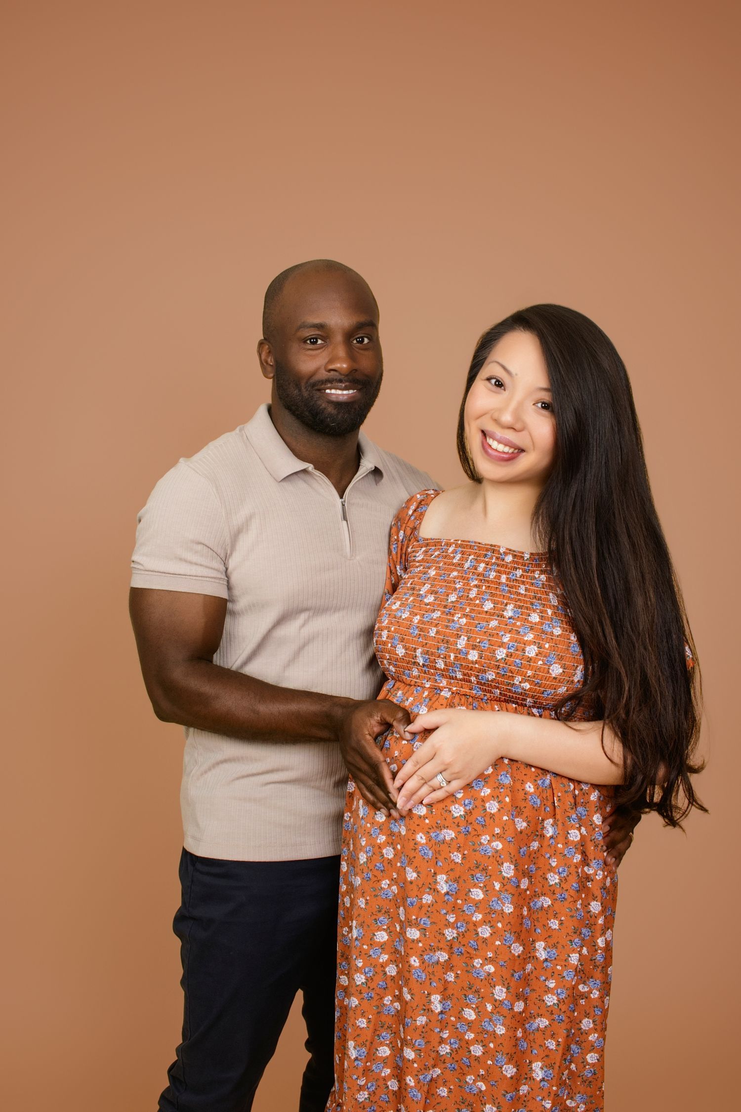 Expectant parents pose lovingly in studio with warm lighting and neutral backdrop.