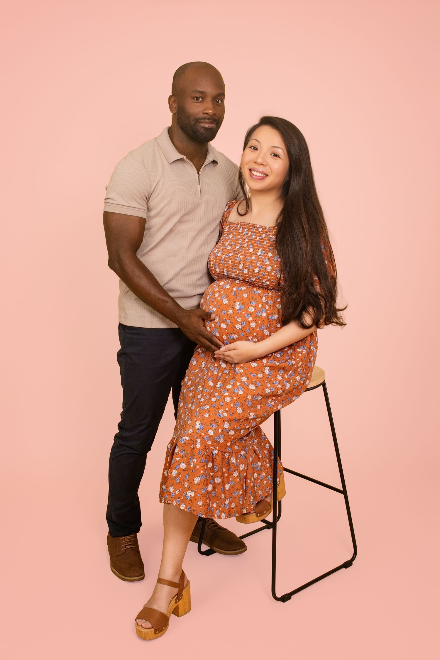 Casual maternity session with couple sitting on black chair against pink backdrop.