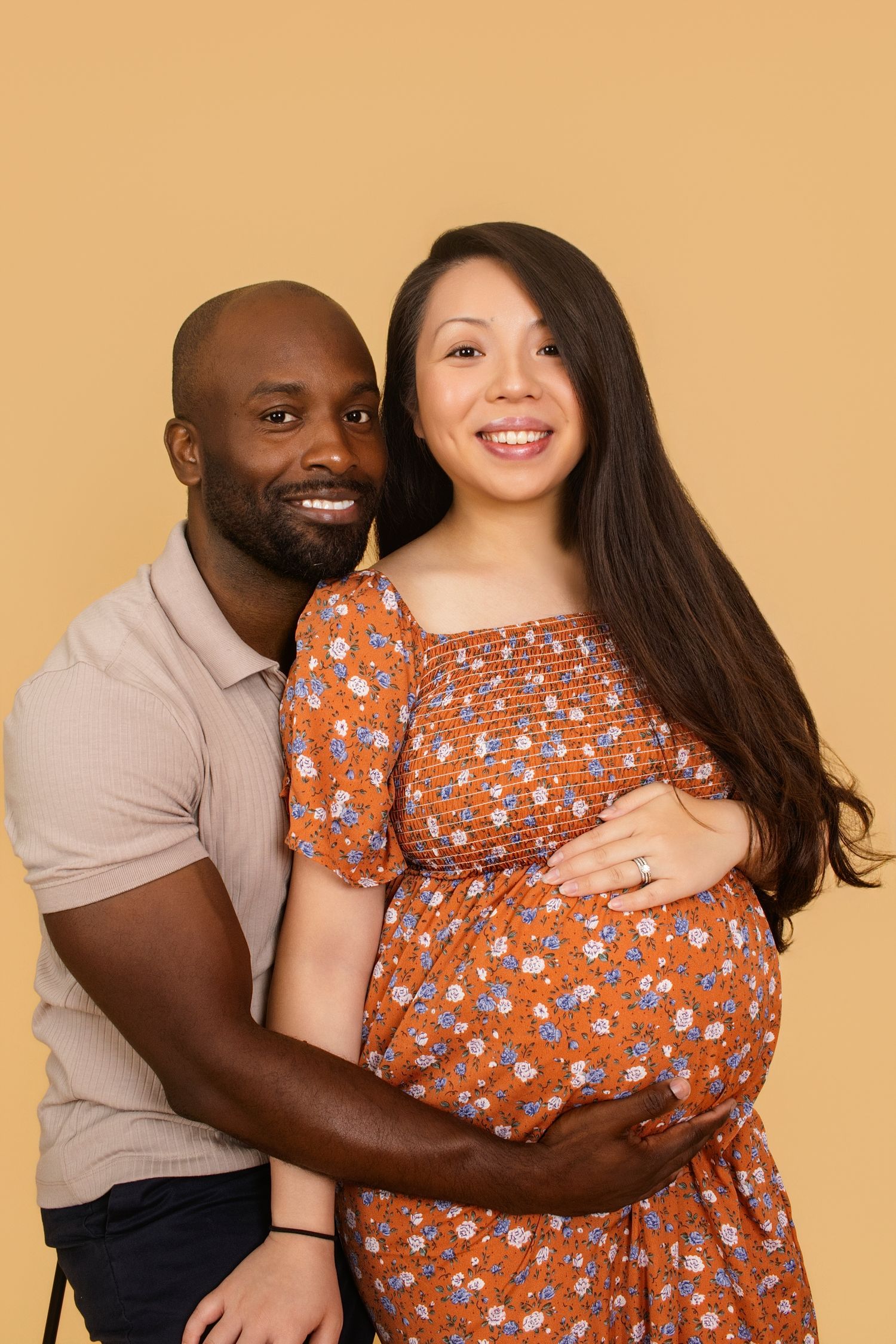 Loving couple's maternity photos with rustic orange floral dress against neutral background.