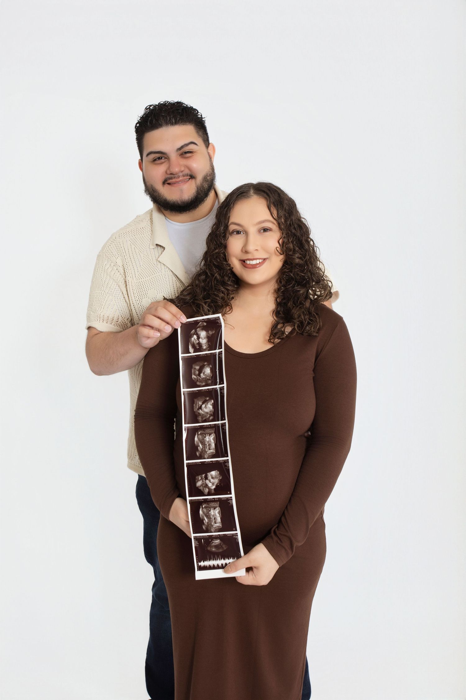 A couple in brown and cream outfits holds up ultrasound photos while smiling.