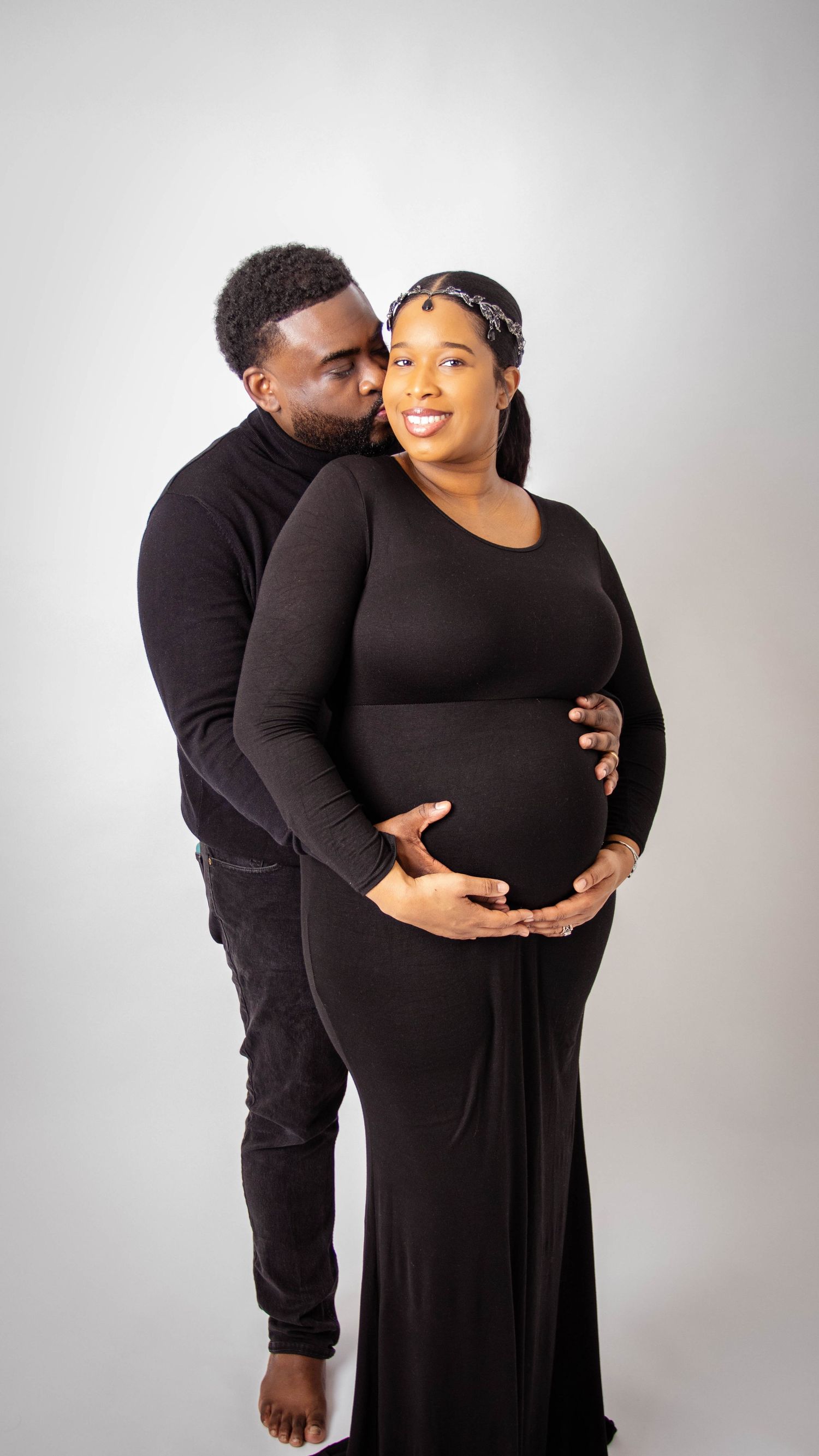 Intimate maternity photo of couple in black outfits embracing against white background.