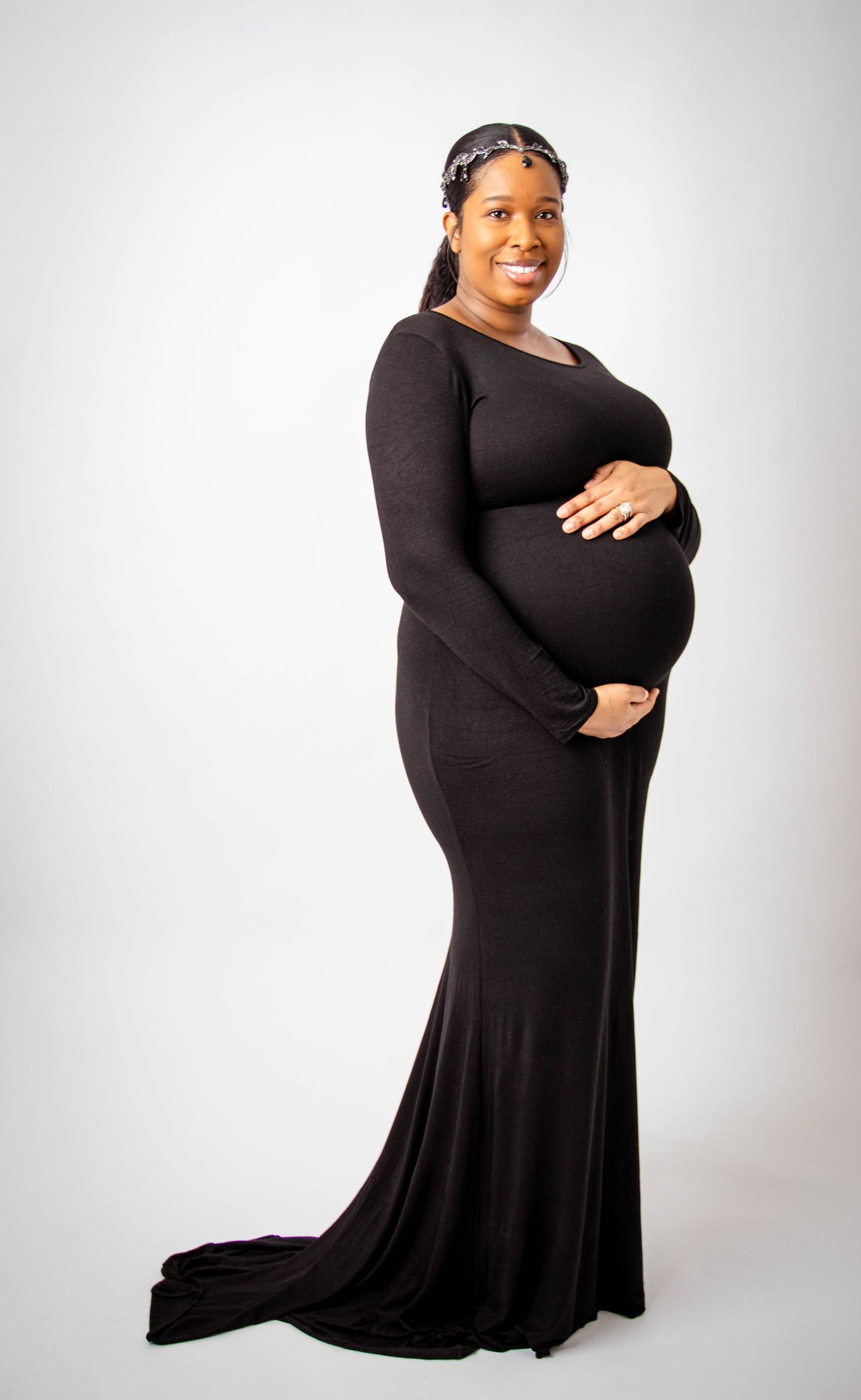 Pregnant woman in flowing black dress poses elegantly against light background.