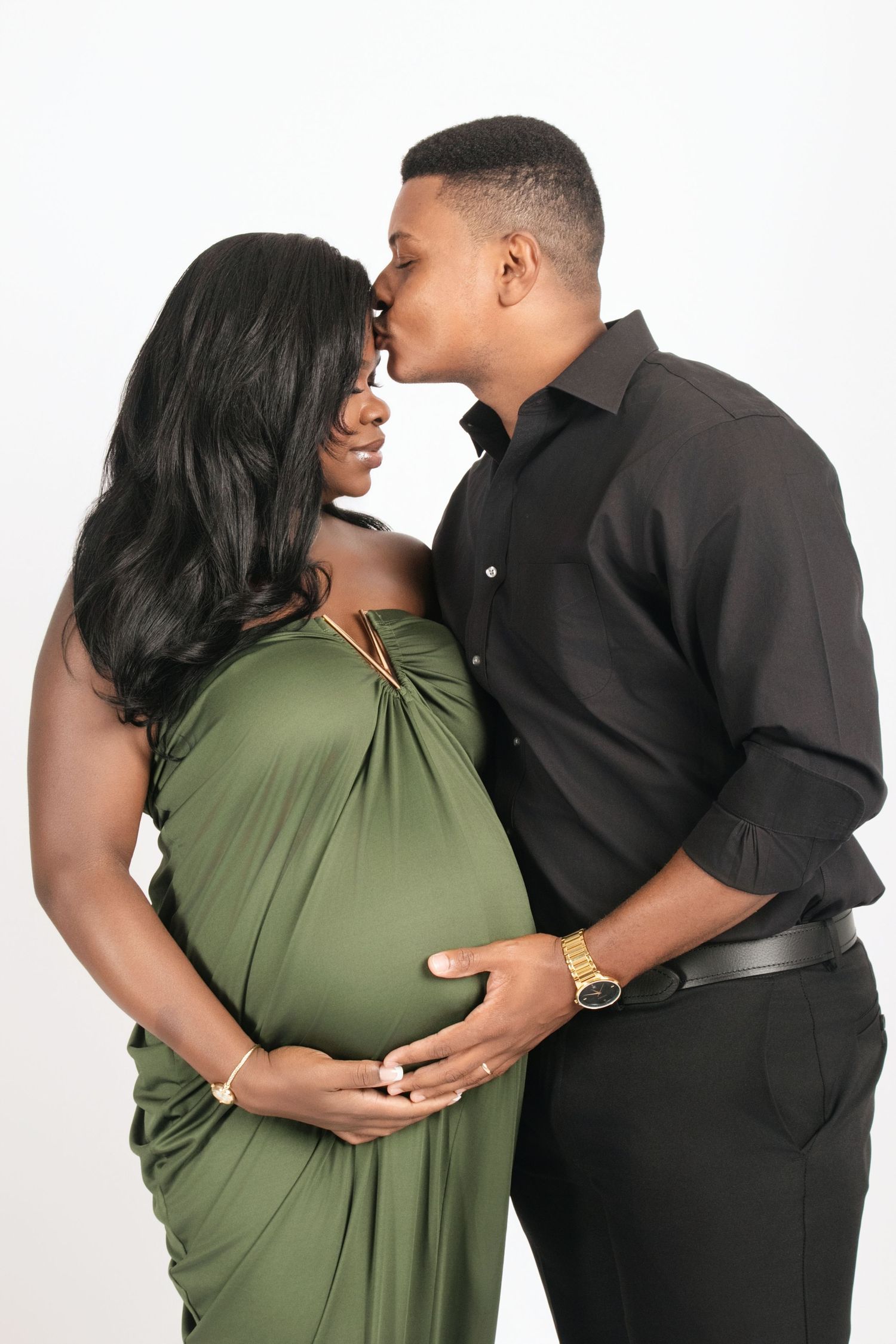 A couple poses together in a romantic maternity photoshoot series wearing green and black outfits against a white backdrop.