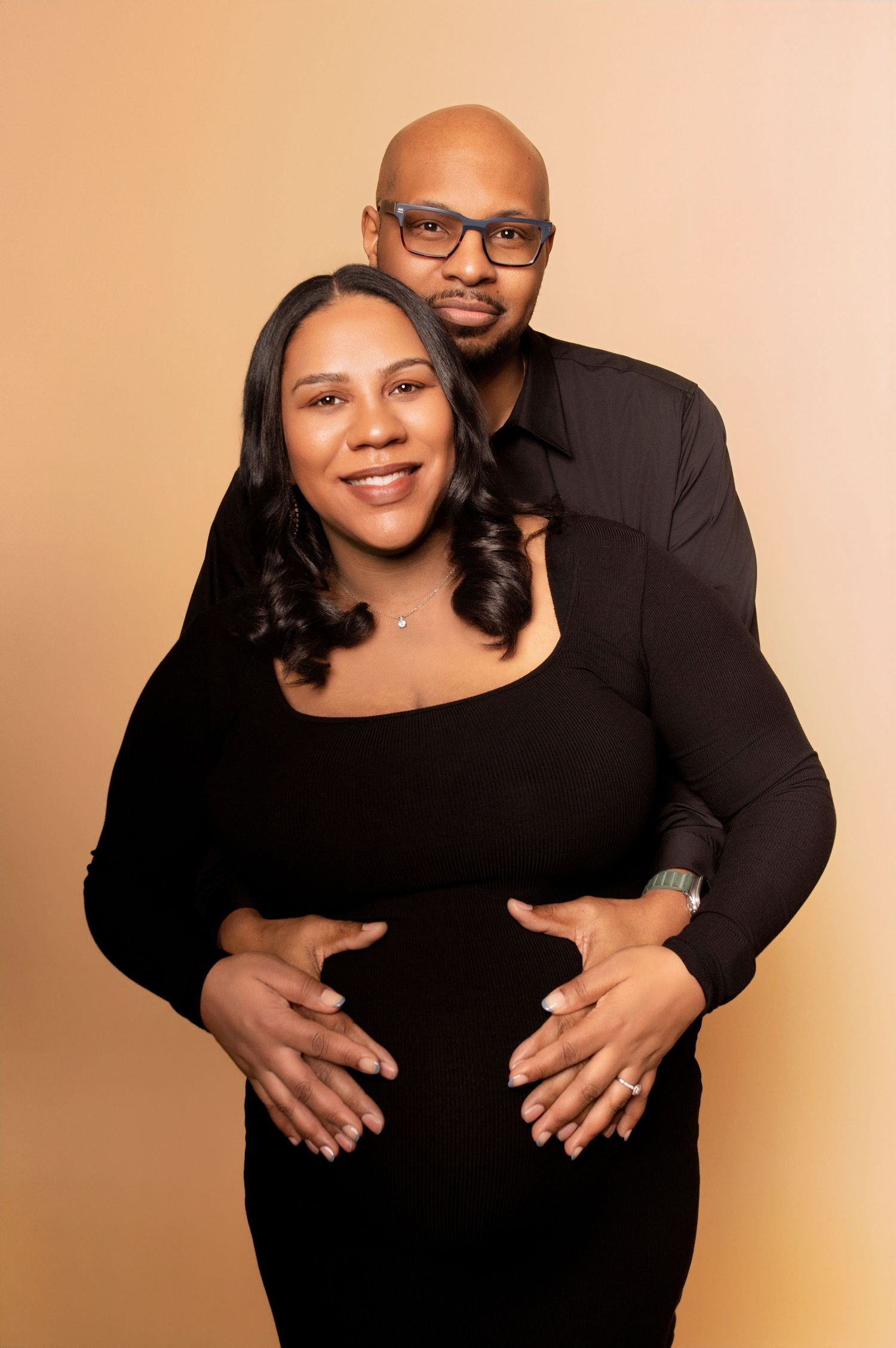 A couple shares tender moments in black outfits during their maternity photoshoot against a peach background.