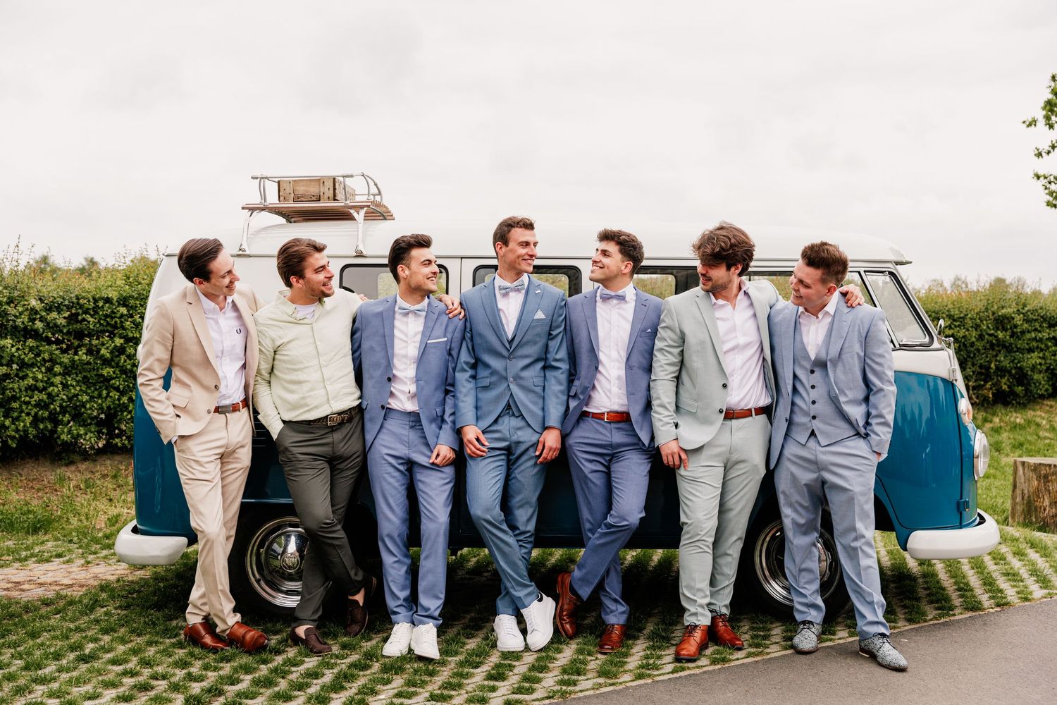 Wedding party members in blue and gray suits sit casually on a stone wall outdoors.