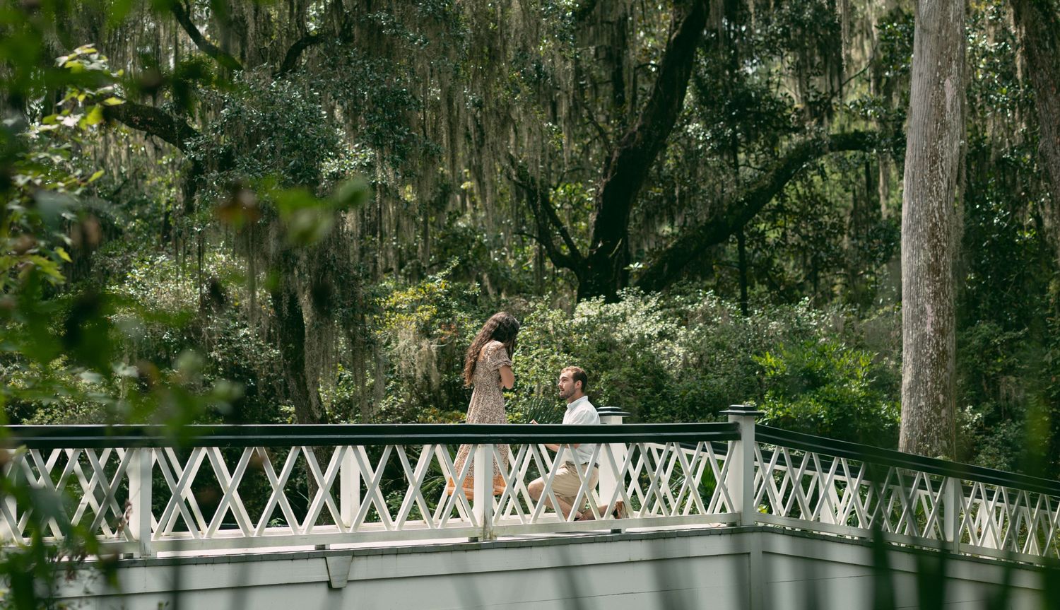A couple stands on a white wooden bridge surrounded by moss-draped trees.