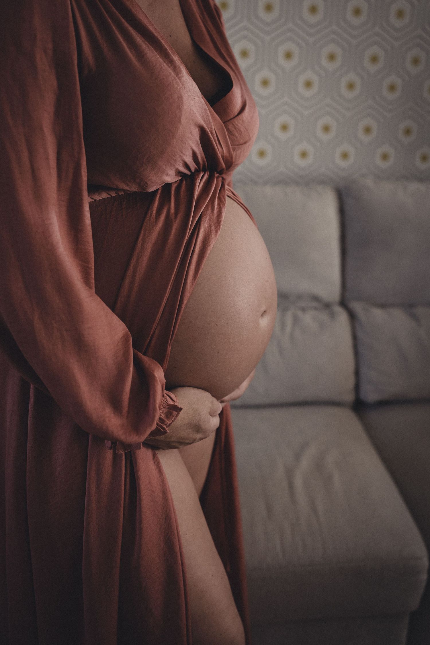 Side view of pregnant belly in burgundy dress against grey couch background.