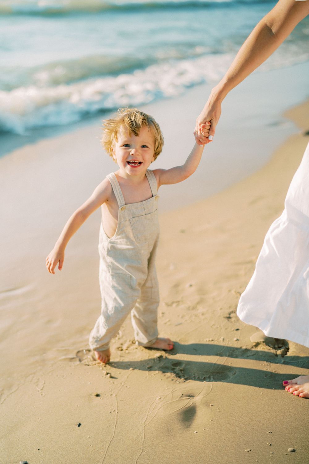 A toddler in overalls walks hand-in-hand with parent along the beach at sunset.