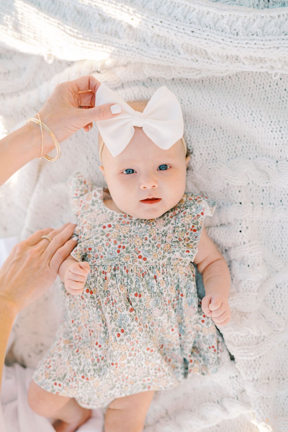 A baby in a floral dress and white bow lying on a lace blanket.