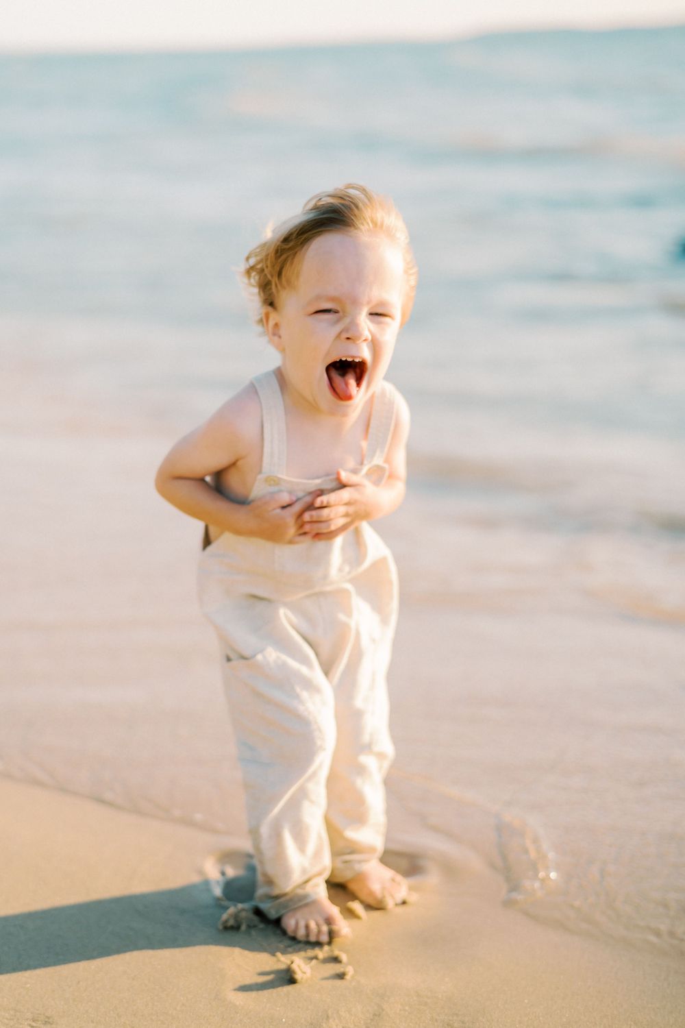Joyful toddler in white overalls laughing while playing on a sandy beach at sunset.