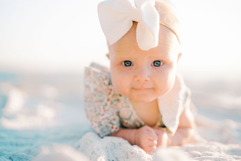 Baby wearing white bow headband lying on beach blanket with gentle smile.