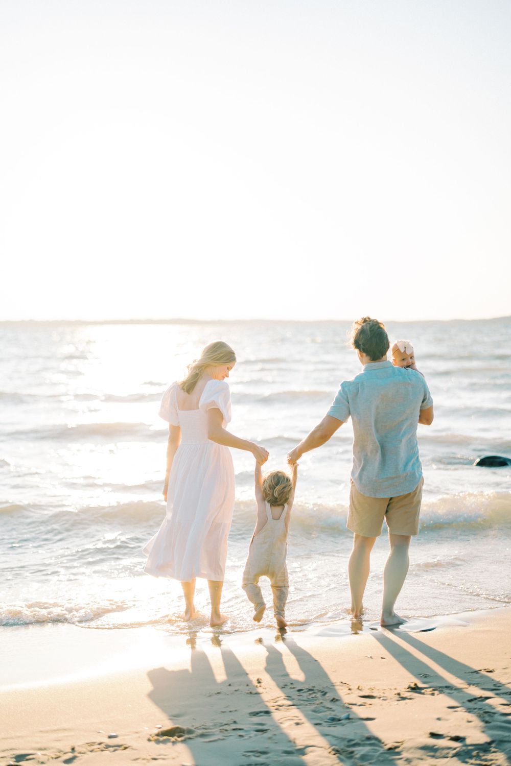 Family of three walking hand in hand along the beach at sunset casting long shadows.