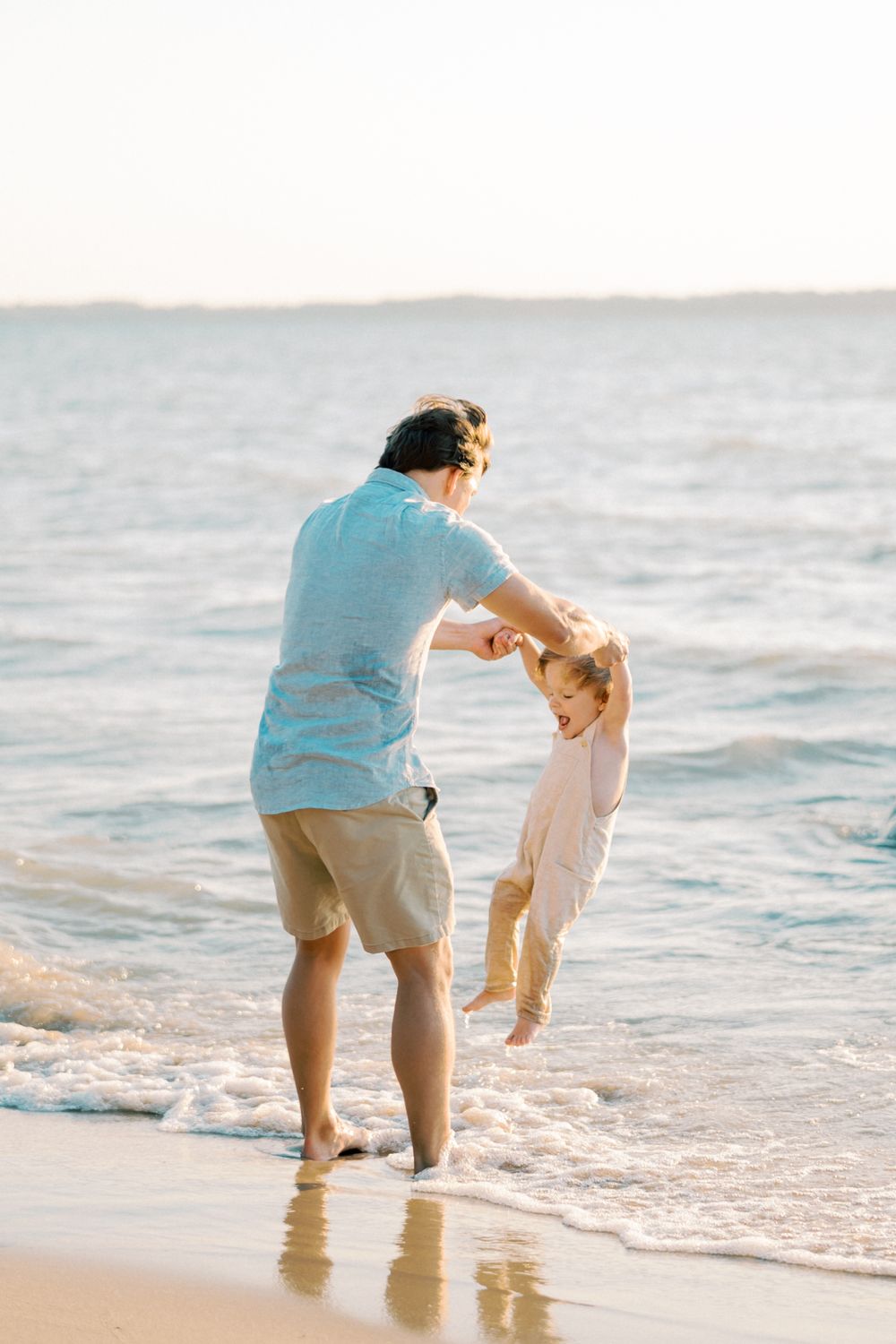 Parent swinging child in white clothing on beach with ocean waves in background.