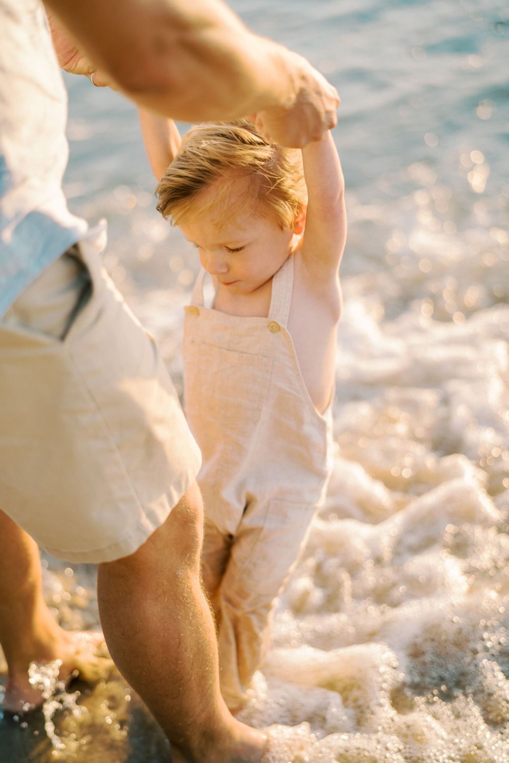 Small child in light colored overalls splashing in ocean waves at sunset.