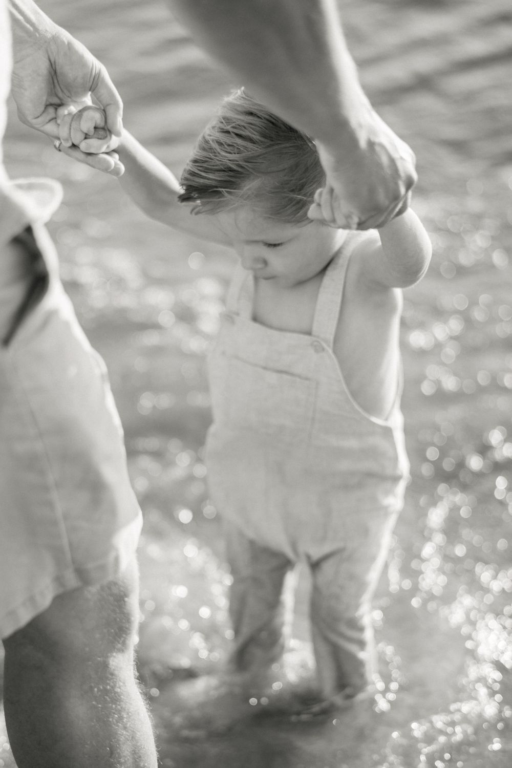 Black and white photo of small child in overalls holding hands with adult near sparkling water.