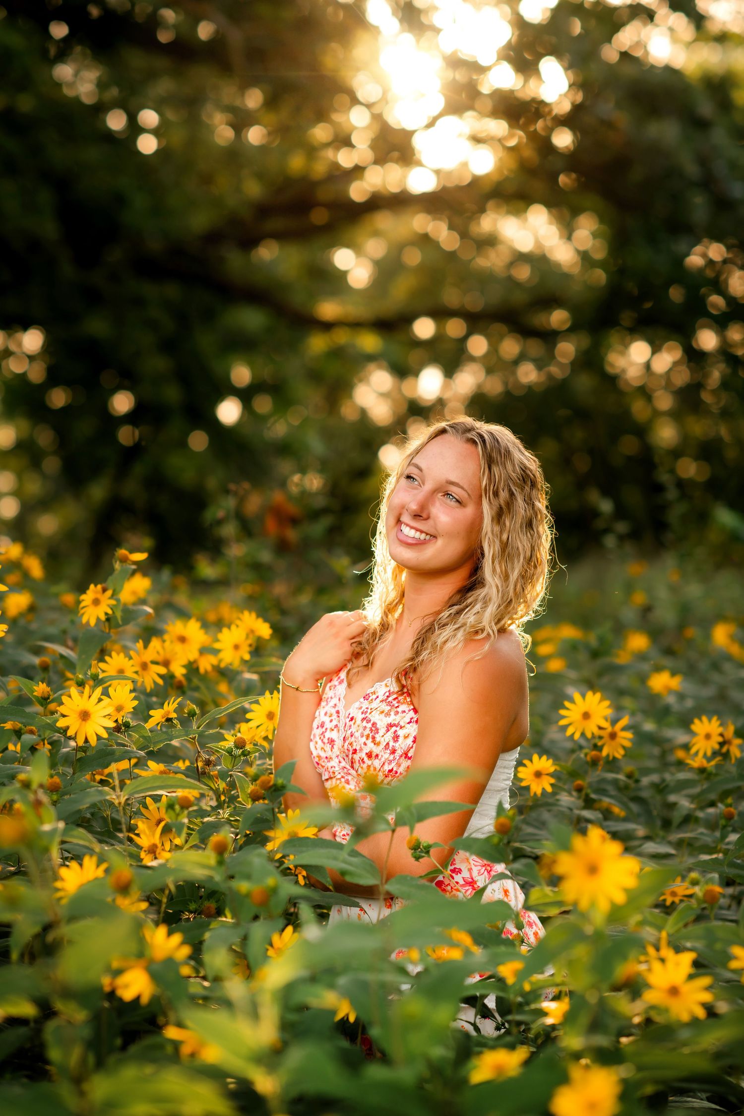 A smiling senior girl stands amidst a field of yellow flowers, with warm sunlight filtering through the foliage.