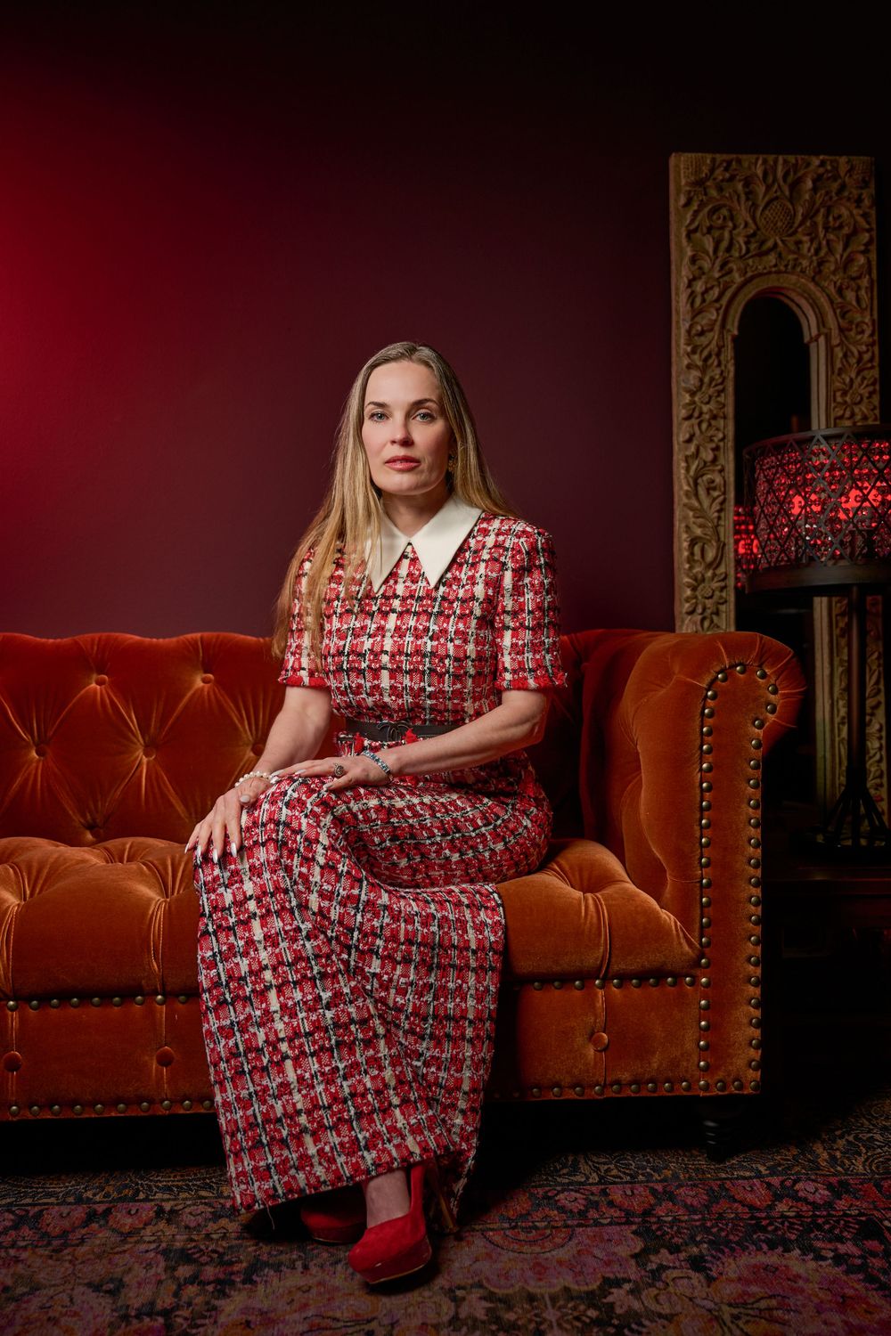 A person in a red and white patterned dress with collar sits elegantly on a tufted orange velvet sofa against dark backdrop.