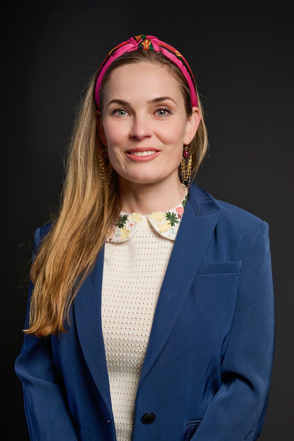 Professional headshot of an individual wearing a navy blazer, white sweater, pink headband and gold earrings against dark background.