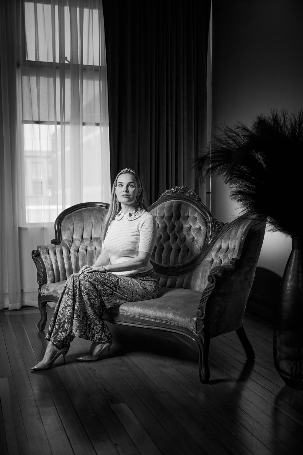 Dramatic black and white photo showing seated figure on vintage tufted sofa near sunlit window with flowing curtains.