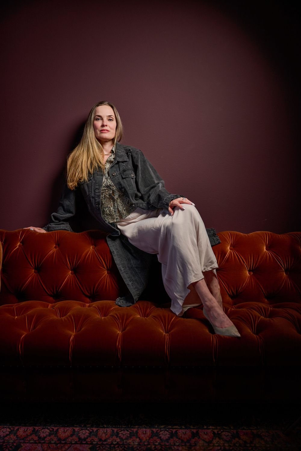 Figure reclining on a luxurious red velvet couch in dramatic lighting wearing denim and casual attire.