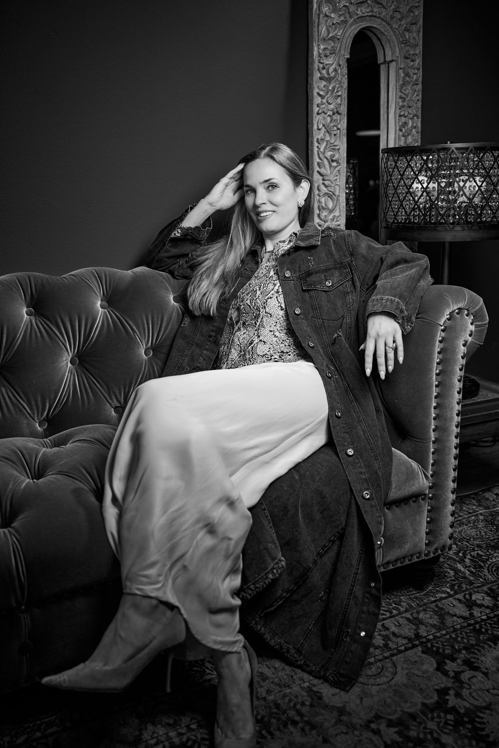 Black and white portrait of someone in denim jacket and floral dress lounging on tufted velvet sofa with decorative lamp.