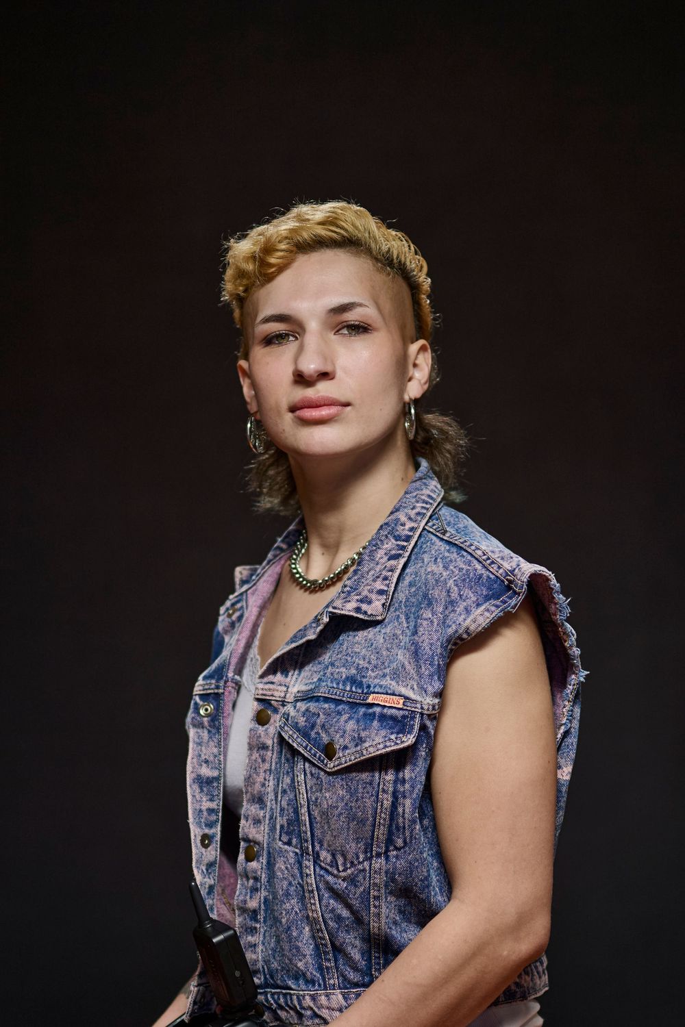 Person in sleeveless distressed denim vest with exaggerated shoulder details poses against dark background.