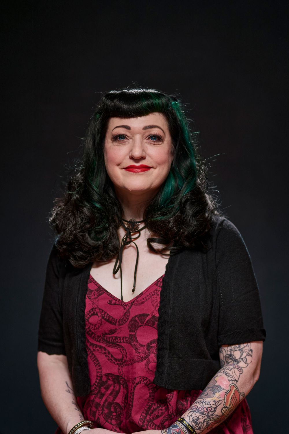 Portrait of a person with dark hair styled in vintage waves, wearing red dress and black cardigan against dark background.