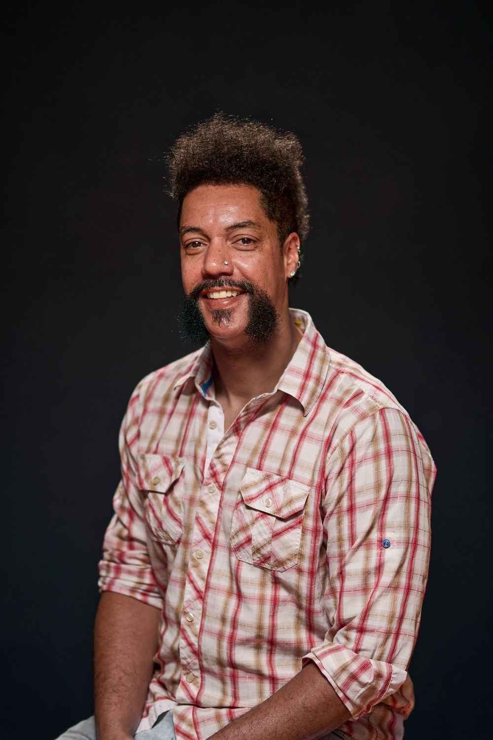 Smiling person wearing a red and white plaid button-up shirt against a dark background.