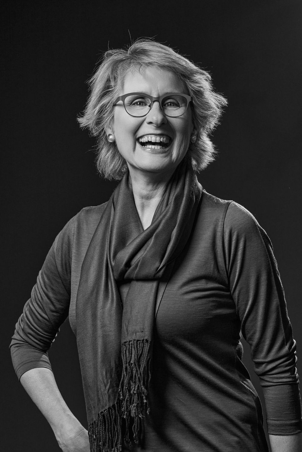 Black and white portrait of a person in glasses and scarf wearing a long-sleeved top, smiling warmly against dark background.