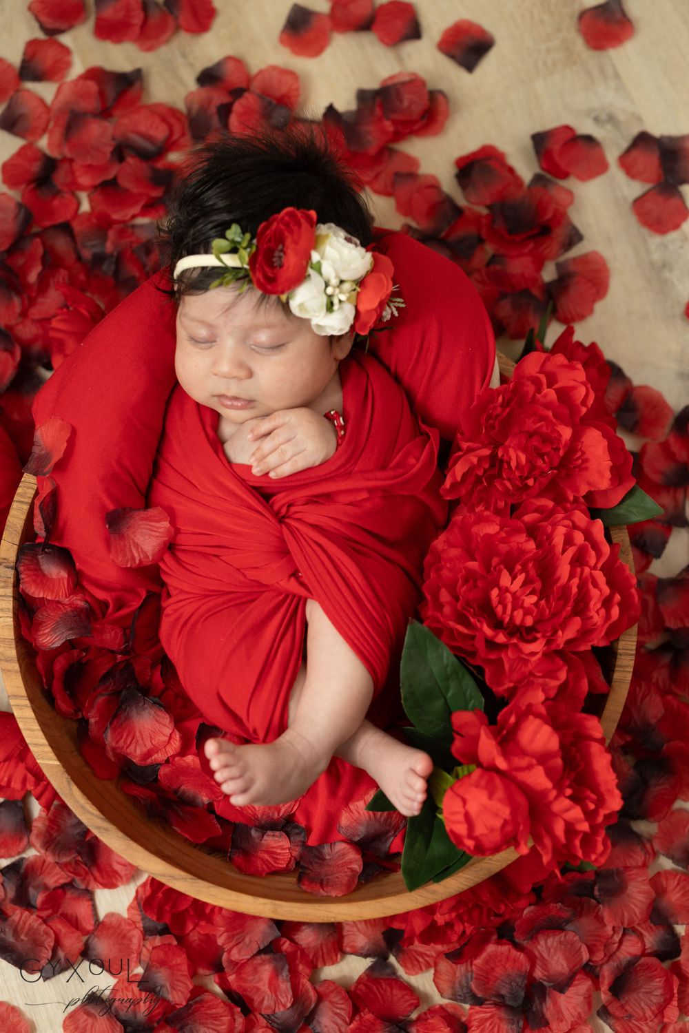 Sleeping newborn wrapped in red fabric surrounded by rose petals in wooden bowl.