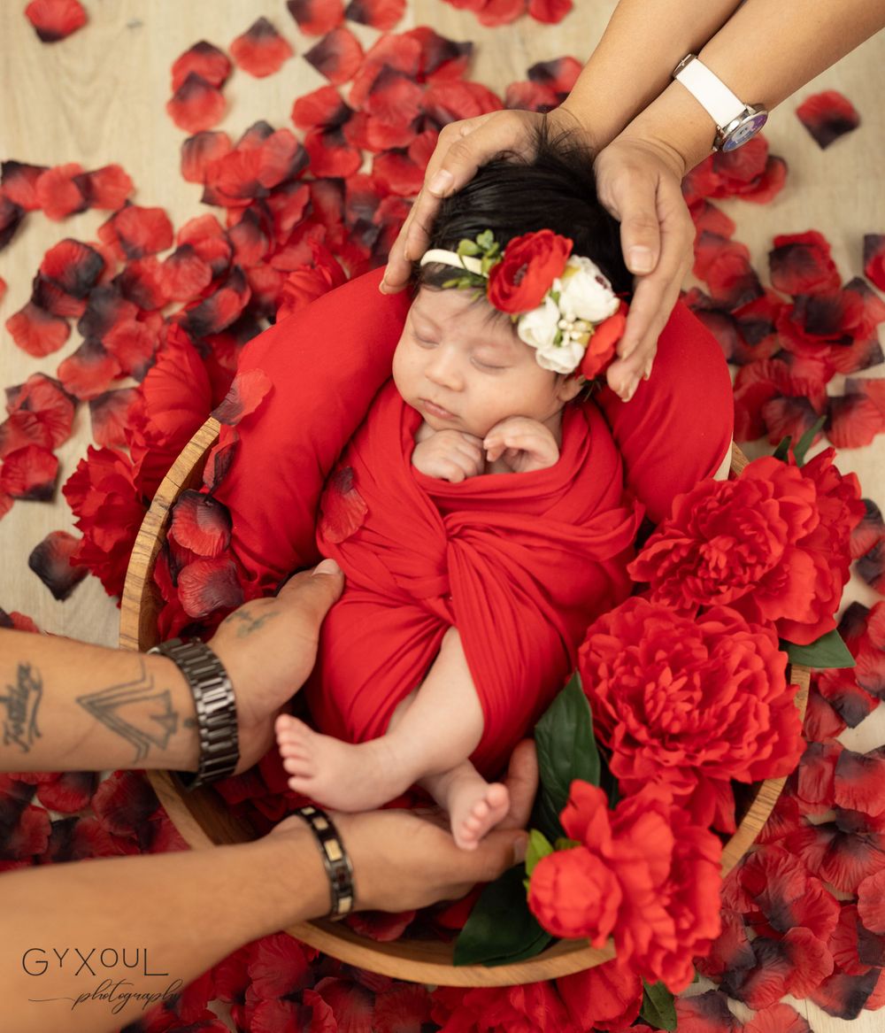 Newborn photography with baby in red wrap surrounded by rose petals and gentle hands.