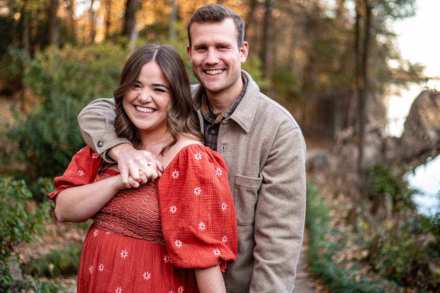 Smiling engaged couple in red dress and grey jacket posing by autumn lakeside.