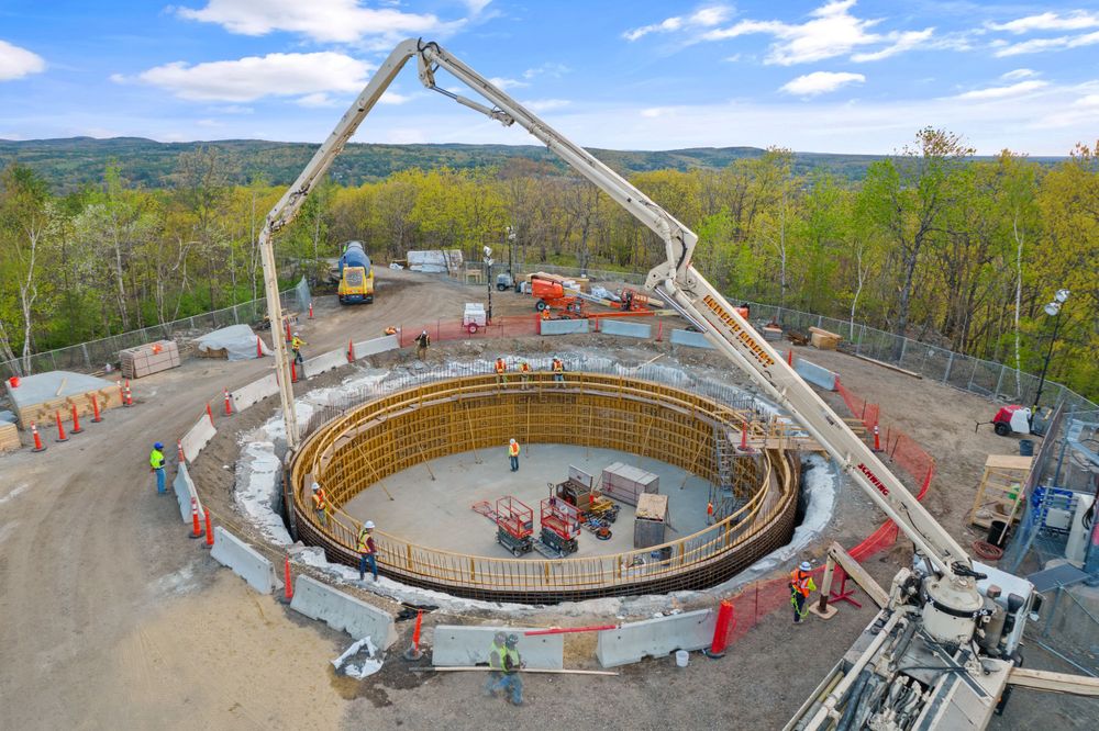 Construction site with concrete pump truck pouring circular foundation surrounded by equipment.