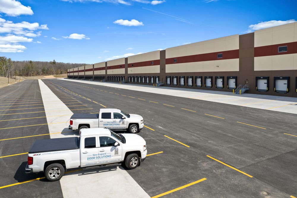 Large industrial warehouse building with multiple loading docks and two white trucks in parking lot.