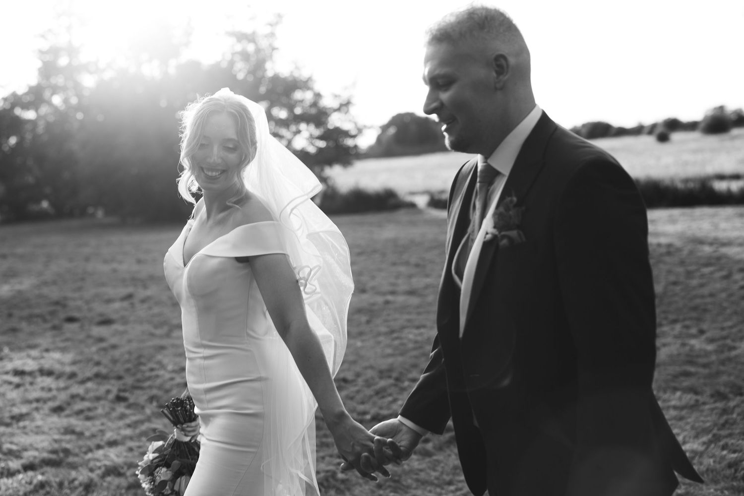 Wedding couple walking hand in hand through a sunlit field.