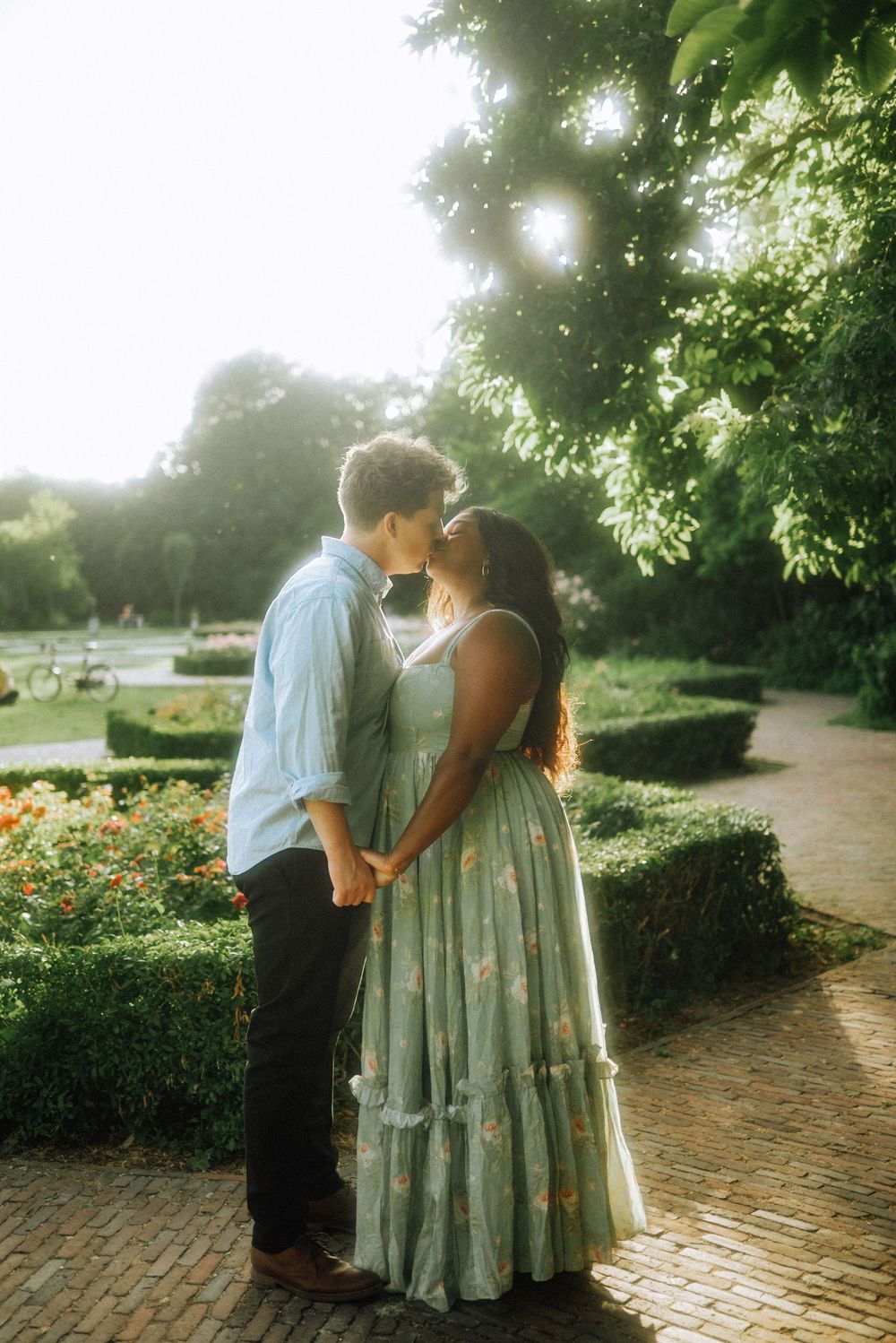 Couple in a sunlit garden share a kiss while holding hands, she in a flowing floral maxi dress, he in a light blue shirt.