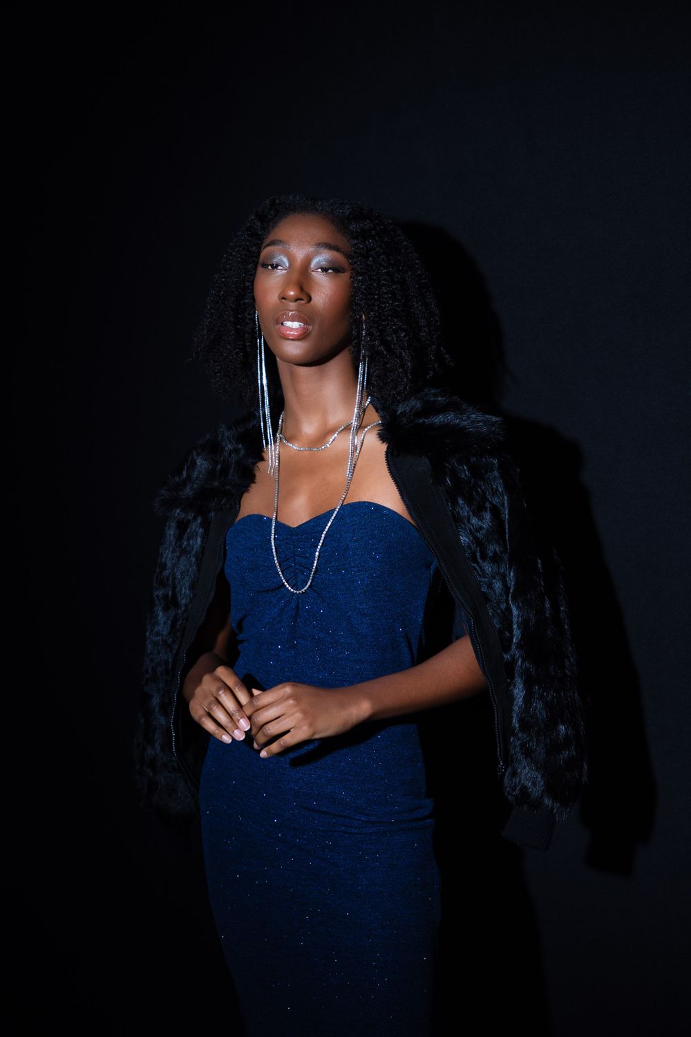 Fashion portrait of model in blue dress and black jacket against dark background.