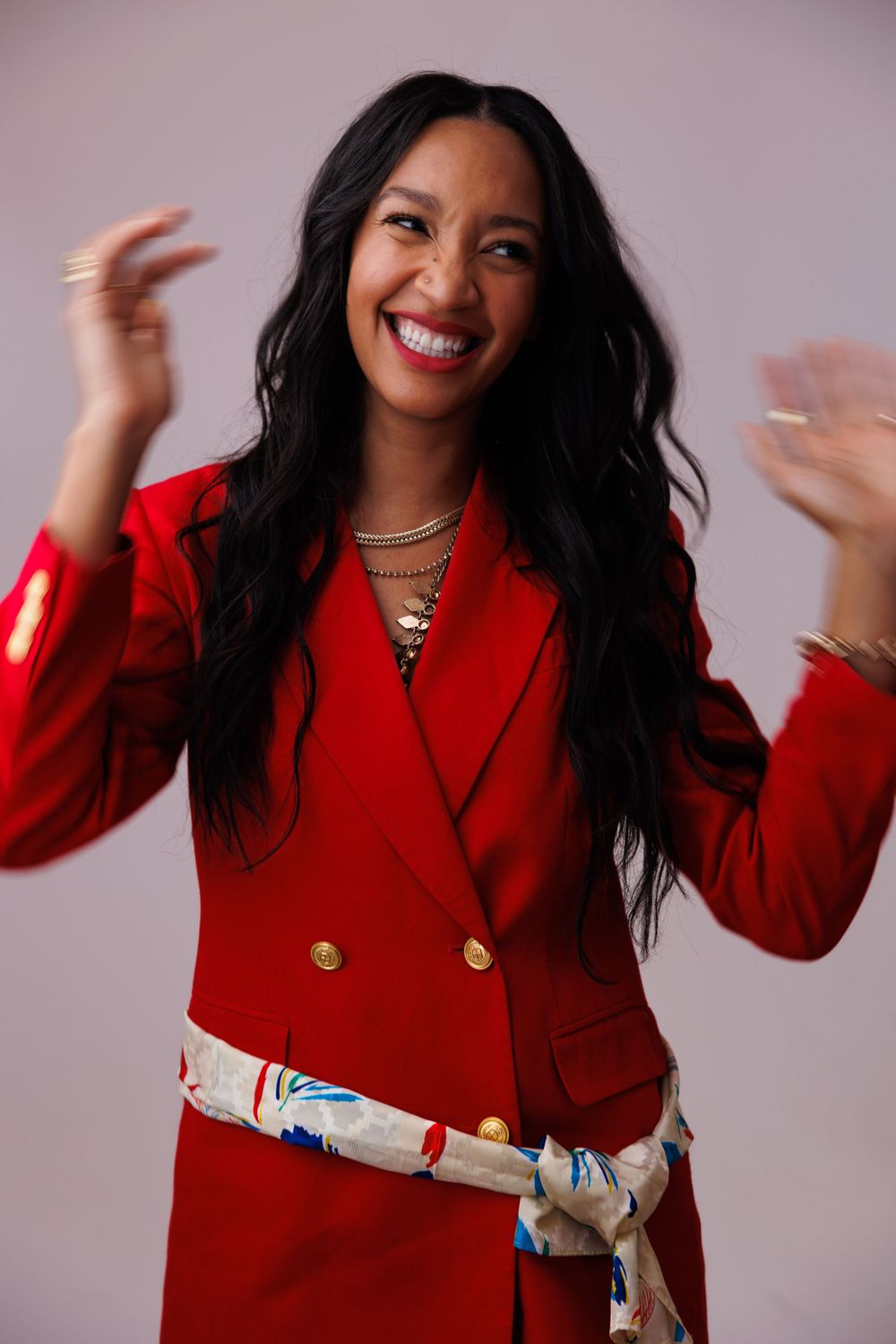 Woman in vibrant red blazer with sash belt smiling and gesturing against pink background.