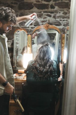 Hairstyling session in progress with hairspray being applied in front of vintage gold-framed mirrors.