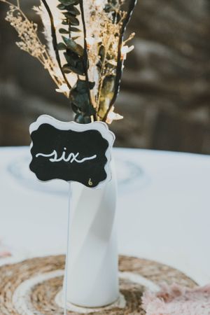White ceramic vase with dried flowers and a black chalkboard tag displaying 'six' on a white tablecloth.