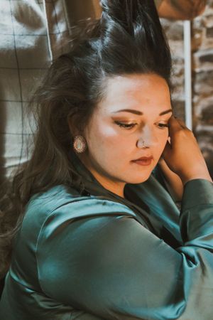 Portrait of a woman in teal satin with dramatic makeup in moody lighting against stone wall.