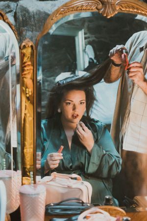 A vintage-style scene of someone applying makeup in front of an ornate gold mirror at a vanity table.