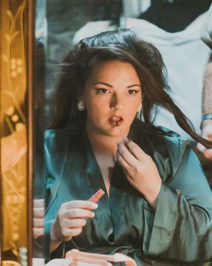 Woman in teal robe applies makeup while looking in mirror with flowing dark hair.
