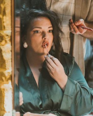 Woman in teal silk blouse having makeup applied near a window with warm lighting.