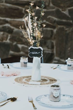 Elegant wedding table setting with white vase centerpiece and delicate dried flowers against stone wall.