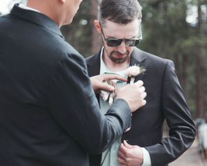 Groomsman helping adjust boutonniere on suit lapel during wedding preparation.
