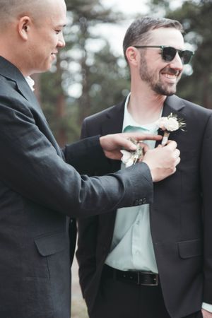 Two men in formal suits at a wedding, with one helping to adjust the other's boutonniere.