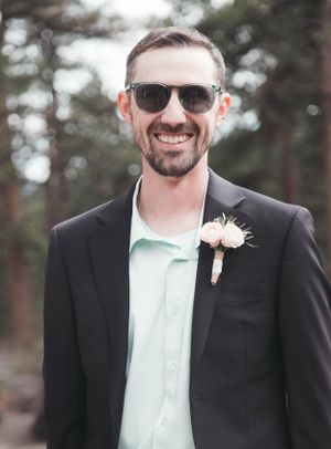 A groom wearing sunglasses and a boutonniere smiles in a wooded setting.